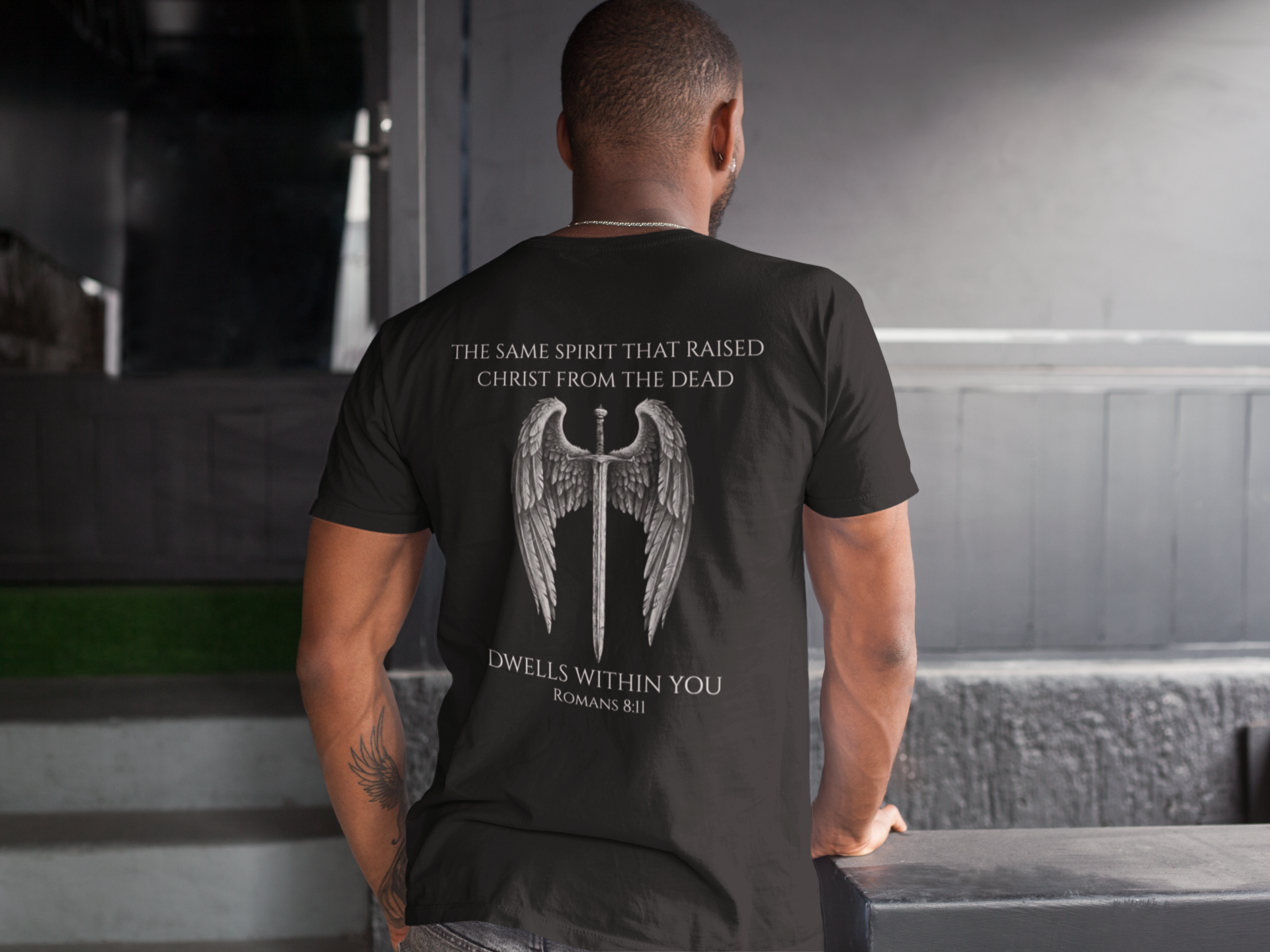 Man wearing a black t-shirt featuring a winged sword graphic with the text “The same Spirit that raised Christ from the dead dwells within you – Romans 8:11” printed on the back.
