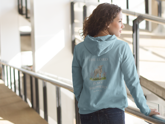 Young woman wearing a light blue Christian hoodie with a lamb illustration and the Bible verse “The Lord Is My Shepherd – Psalm 23” on the back, standing indoors by a railing with natural light.
